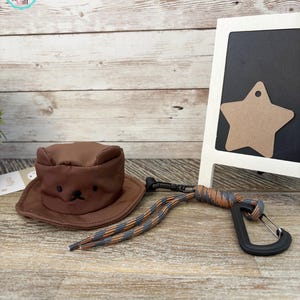 May include: A brown pet hat with a bear face design, a leash with a black carabiner, and a decorative star-shaped tag. The hat is made of a soft material and has a wide brim. The leash is gray, orange, and black.
