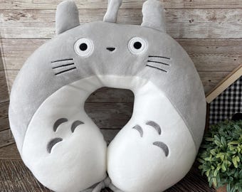 May include: Gray and white plush travel pillow shaped like a cartoon cat with a white face and gray ears. The pillow has a hole in the middle for the neck and a gray tie to secure it.