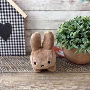 May include: A plush brown bunny keychain with black eyes and a small black cross for a mouth. The bunny is standing on a wooden surface. A small house with a black and white checkered pattern is in the background.