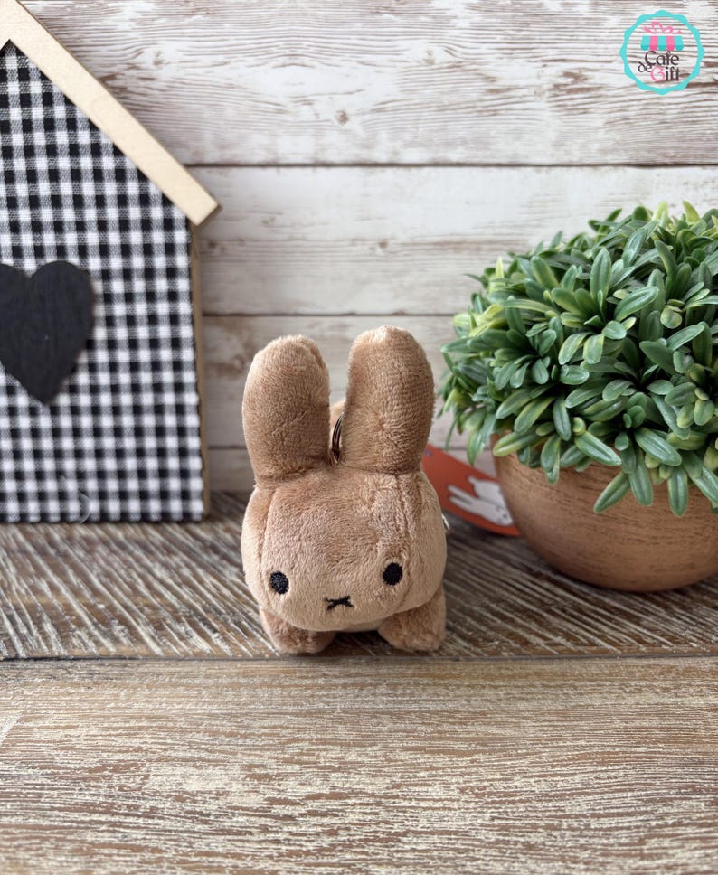 May include: A plush brown bunny keychain with black eyes and a small black cross for a mouth. The bunny is standing on a wooden surface. A small house with a black and white checkered pattern is in the background.