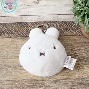 May include: A white plush Miffy keychain with a round body, two small ears, and a silver-colored metal clasp. The face features small brown eyes and a stitched brown cross for a mouth. A small tag with the word "Miffy" is attached.