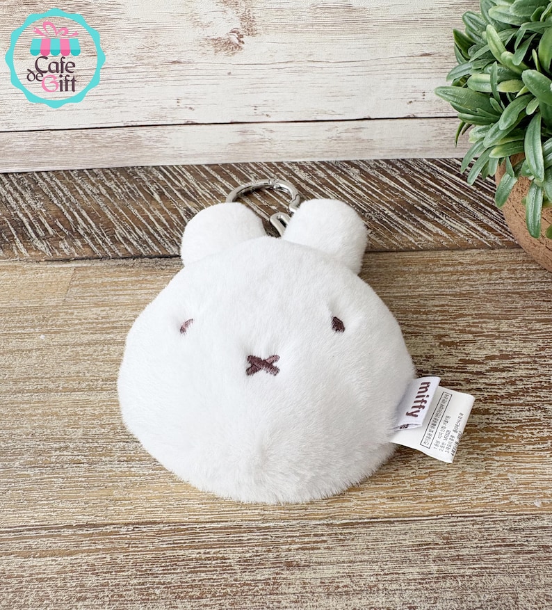 May include: A white plush Miffy keychain with a round body, two small ears, and a silver-colored metal clasp. The face features small brown eyes and a stitched brown cross for a mouth. A small tag with the word "Miffy" is attached.