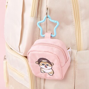 May include: A pink mini backpack keychain with a cartoon dog wearing a hat embroidered on the front. The keychain has a blue star-shaped clip and is attached to a beige backpack. The mini backpack has a zipper closure.