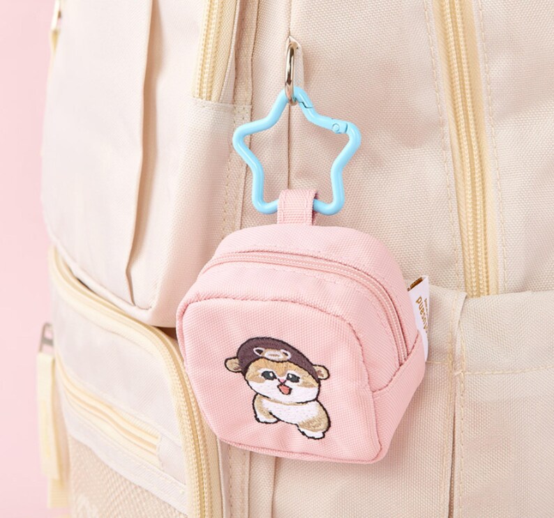 May include: A pink mini backpack keychain with a cartoon dog wearing a hat embroidered on the front. The keychain has a blue star-shaped clip and is attached to a beige backpack. The mini backpack has a zipper closure.
