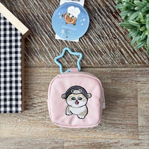May include: A light pink square coin purse with a cartoon cat wearing a hat. The purse has a blue star-shaped clasp. A round tag with a cartoon character and the word "mofusand" is attached.