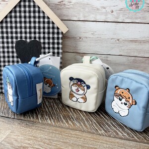 May include: Three small, square backpacks in blue and cream colors, each featuring an embroidered cat design. The backpacks have zippers and small handles. A wooden house and a black heart are in the background.