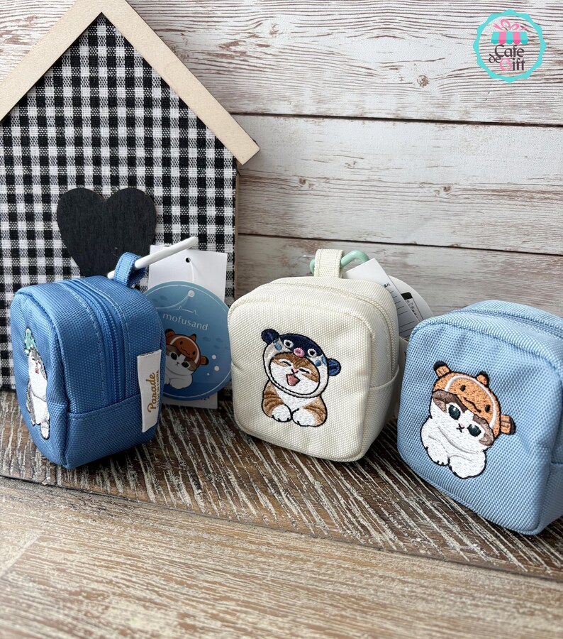 May include: Three small, square backpacks in blue and cream colors, each featuring an embroidered cat design. The backpacks have zippers and small handles. A wooden house and a black heart are in the background.