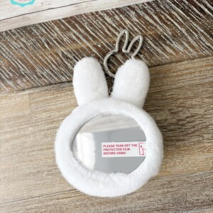 May include: A white, plush bunny-shaped compact mirror with a silver-tone bunny ear keychain. The mirror has a protective film with the text "PLEASE TEAR OFF THE PROTECTIVE FILM BEFORE USING". The Cafe de Gift logo is in the upper left corner.