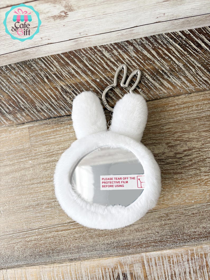 May include: A white, plush bunny-shaped compact mirror with a silver-tone bunny ear keychain. The mirror has a protective film with the text "PLEASE TEAR OFF THE PROTECTIVE FILM BEFORE USING". The Cafe de Gift logo is in the upper left corner.