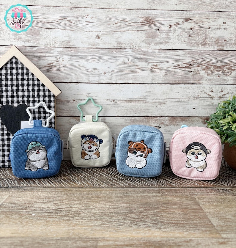 May include: Four small, square pouches in blue, white, light blue, and pink, each with a different embroidered cat design. The pouches have a zipper closure and a star-shaped or circular clip. The background is a wooden surface.