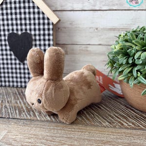 May include: A plush brown bunny toy with black bead eyes. The bunny is lying down on a wooden surface. A small decorative house with a black and white checkered pattern is in the background.