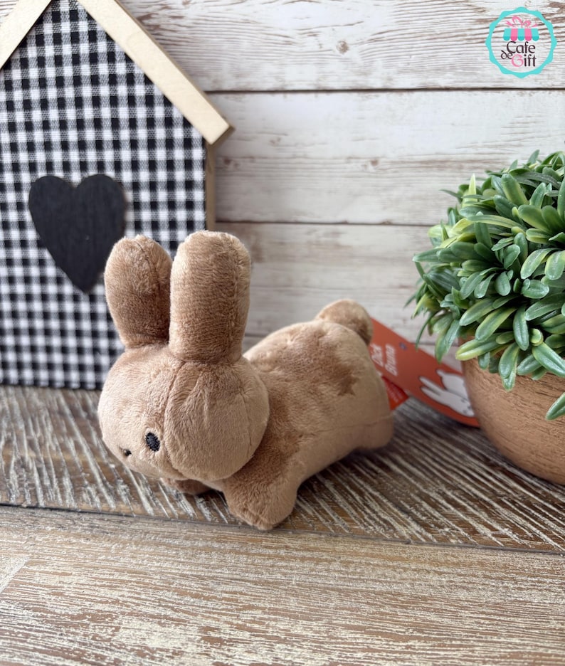 May include: A plush brown bunny toy with black bead eyes. The bunny is lying down on a wooden surface. A small decorative house with a black and white checkered pattern is in the background.