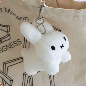 May include: A white plush bunny keychain with black embroidered eyes and a cross-shaped mouth. The keychain is attached to a silver metal clasp and ring, hanging from a beige tote bag.