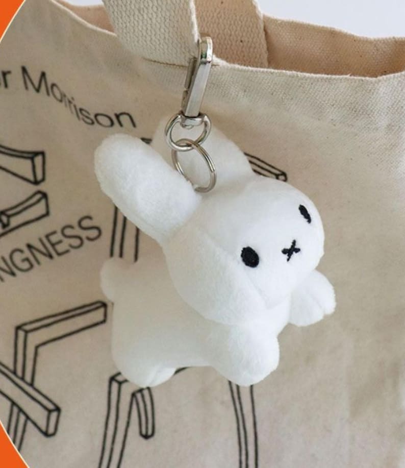 May include: A white plush bunny keychain with black embroidered eyes and a cross-shaped mouth. The keychain is attached to a silver metal clasp and ring, hanging from a beige tote bag.