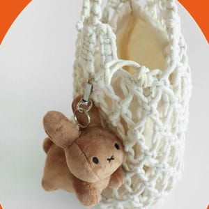 May include: A white macrame bag with a beige interior, holding a brown plush bunny keychain. The bunny has black eyes and a small black cross for a mouth. The bag has a silver clasp.