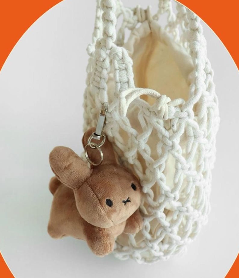 May include: A white macrame bag with a beige interior, holding a brown plush bunny keychain. The bunny has black eyes and a small black cross for a mouth. The bag has a silver clasp.