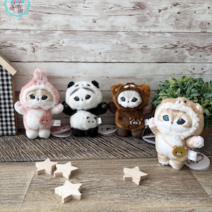 May include: Four plush toys in animal costumes: a pink bunny, a panda, a brown bear, and a tan cat. Each toy has large eyes and is wearing a costume. Wooden star decorations are scattered in front of the toys.