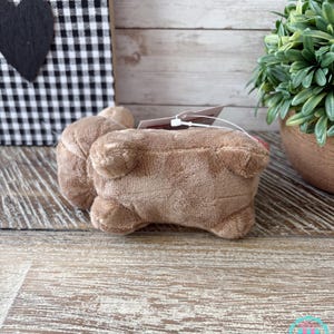 May include: A plush, light brown stuffed animal in the shape of a pig, lying on its belly. The pig has small ears and a tag attached. The background includes a black and white checkered pattern and a small potted plant.
