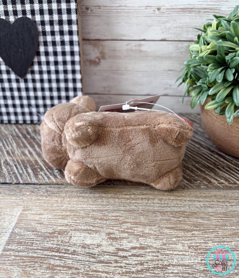 May include: A plush, light brown stuffed animal in the shape of a pig, lying on its belly. The pig has small ears and a tag attached. The background includes a black and white checkered pattern and a small potted plant.