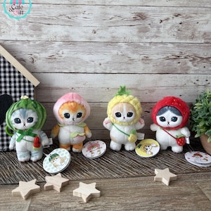 May include: Four plush cat dolls dressed as fruits: watermelon, peach, pineapple, and strawberry. Each doll has a matching fruit-themed hat and a small accessory. Wooden star cutouts and round coasters with cat illustrations are also present.