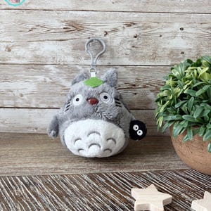 May include: A gray and white plush Totoro keychain with a green leaf on its head and a small black soot sprite attached to its side. The keychain is hanging from a silver clip.