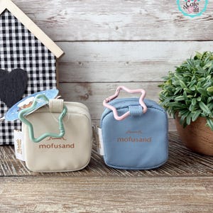 May include: Two small, square pouches in beige and blue, each with a star-shaped clasp. The beige pouch has a green clasp, and the blue pouch has a pink clasp. Both pouches have the word "mofusand" printed on them.