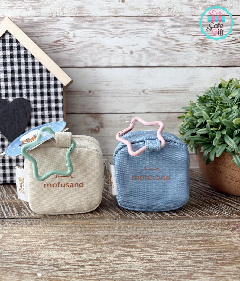 May include: Two small, square pouches in beige and blue, each with a star-shaped clasp. The beige pouch has a green clasp, and the blue pouch has a pink clasp. Both pouches have the word "mofusand" printed on them.