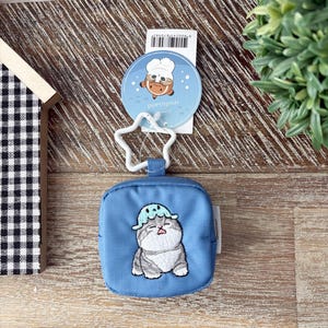 May include: A blue square pouch with a cat design wearing a hat. The cat is gray and white with a blue hat. Attached is a white star-shaped keychain and a round tag with a cartoon character and the word "mousand".
