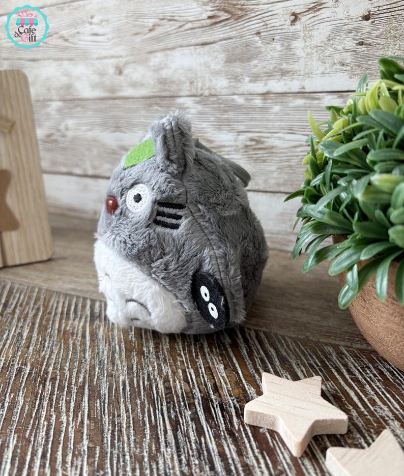 Puede incluir: Un juguete de peluche gris con la barriga blanca y ojos negros. El juguete tiene una oreja verde y un patr&oacute;n de rayas blancas y negras en la espalda. El juguete est&aacute; sentado sobre una superficie de madera.