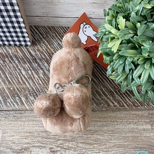 May include: A plush, light brown keychain with two small pom-poms and a silver-colored clasp. The keychain is attached to a larger, rounded plush shape. A small orange card with a white rabbit illustration is visible in the background.