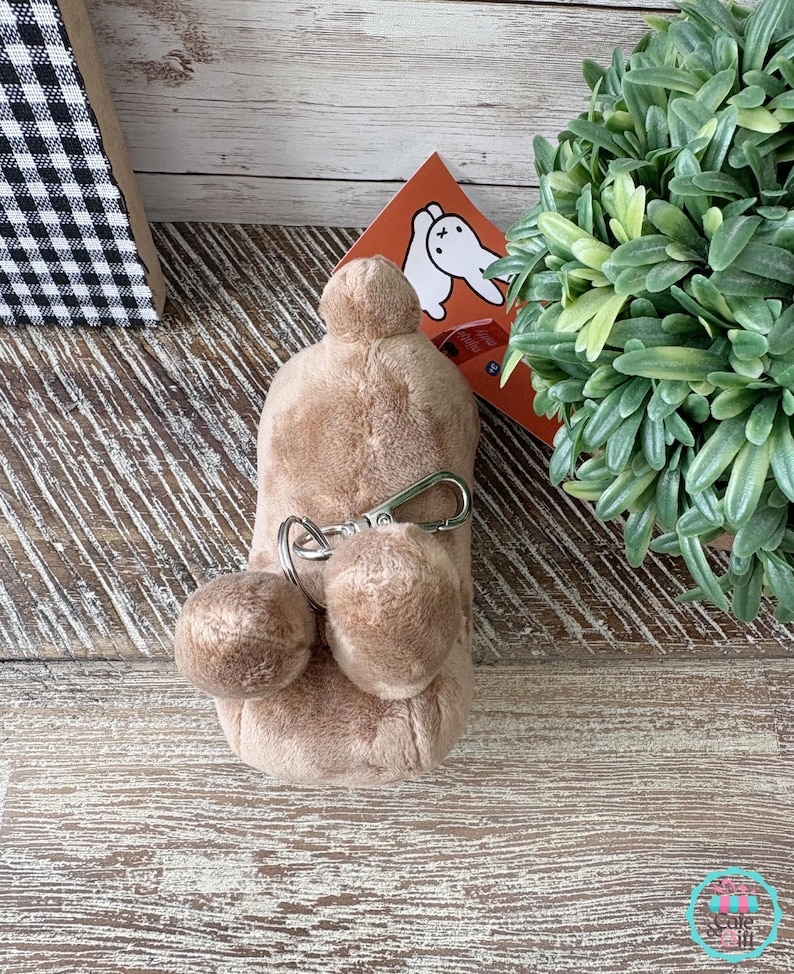 May include: A plush, light brown keychain with two small pom-poms and a silver-colored clasp. The keychain is attached to a larger, rounded plush shape. A small orange card with a white rabbit illustration is visible in the background.