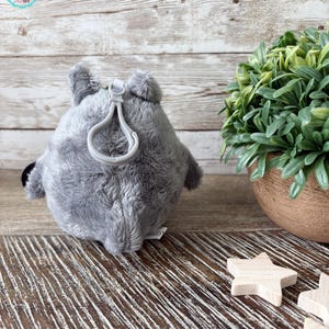 Puede incluir: Llavero de peluche gris en forma de animal redondo con un lazo para sujetarlo a una bolsa o llaves. El llavero est&aacute; sobre una superficie de madera con una planta verde en una maceta en el fondo.