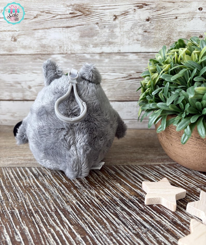Puede incluir: Llavero de peluche gris en forma de animal redondo con un lazo para sujetarlo a una bolsa o llaves. El llavero est&aacute; sobre una superficie de madera con una planta verde en una maceta en el fondo.