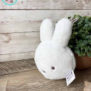 May include: A white plush bunny toy with long ears and a small, round body. The bunny has a small, dark pink nose and eyes. A silver keychain is attached to the top of the bunny. The toy is on a wooden surface.