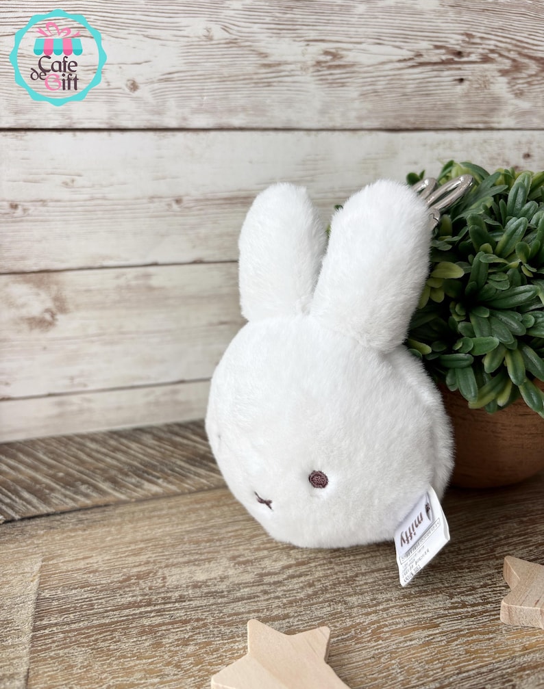 May include: A white plush bunny toy with long ears and a small, round body. The bunny has a small, dark pink nose and eyes. A silver keychain is attached to the top of the bunny. The toy is on a wooden surface.