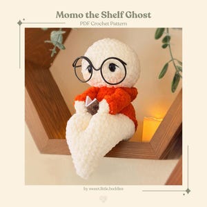 Może przedstawiać: Biała szydełkowa maskotka ducha o nazwie Momo the Shelf Ghost, ubrana w jaskrawopomarańczowy sweter z kapturem i duże czarne okrągłe okulary. Duch siedzi i trzyma małą otwartą książkę. Tekst brzmi Momo the Shelf Ghost PDF Crochet Pattern.