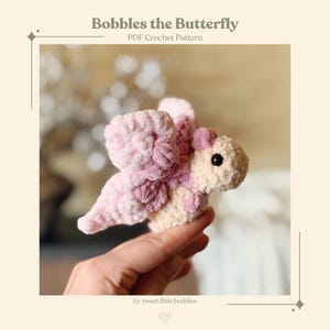 Puede incluir: Una mariposa de ganchillo hecha a mano en tonos rosa y crema. La mariposa tiene alas grandes y esponjosas y un cuerpo suave y texturizado. El texto "Bobbles the Butterfly PDF Crochet Pattern" se muestra en la parte superior de la imagen.