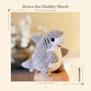 Puede incluir: Un peluche de tiburón tejido a mano en gris y blanco. El tiburón tiene un cuerpo redondo, aletas y una cara sonriente con ojos negros. El texto "Bruce the Chubby Shark" y "PDF Crochet Pattern" son visibles.