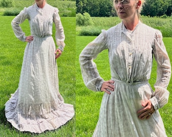 May include: A white cotton dress with a ruffled hem and long sleeves. The dress has a gathered bodice and a fitted waist. The dress is photographed on a person standing in a field.