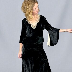 Puede incluir: Un vestido de terciopelo negro con escote en V y mangas acampanadas. El escote est&aacute; adornado con un ribete decorativo de color claro. El vestido parece ser de estilo vintage o retro, con una falda larga y un corpi&ntilde;o ajustado. Las mangas tienen un forro crema en contraste.
