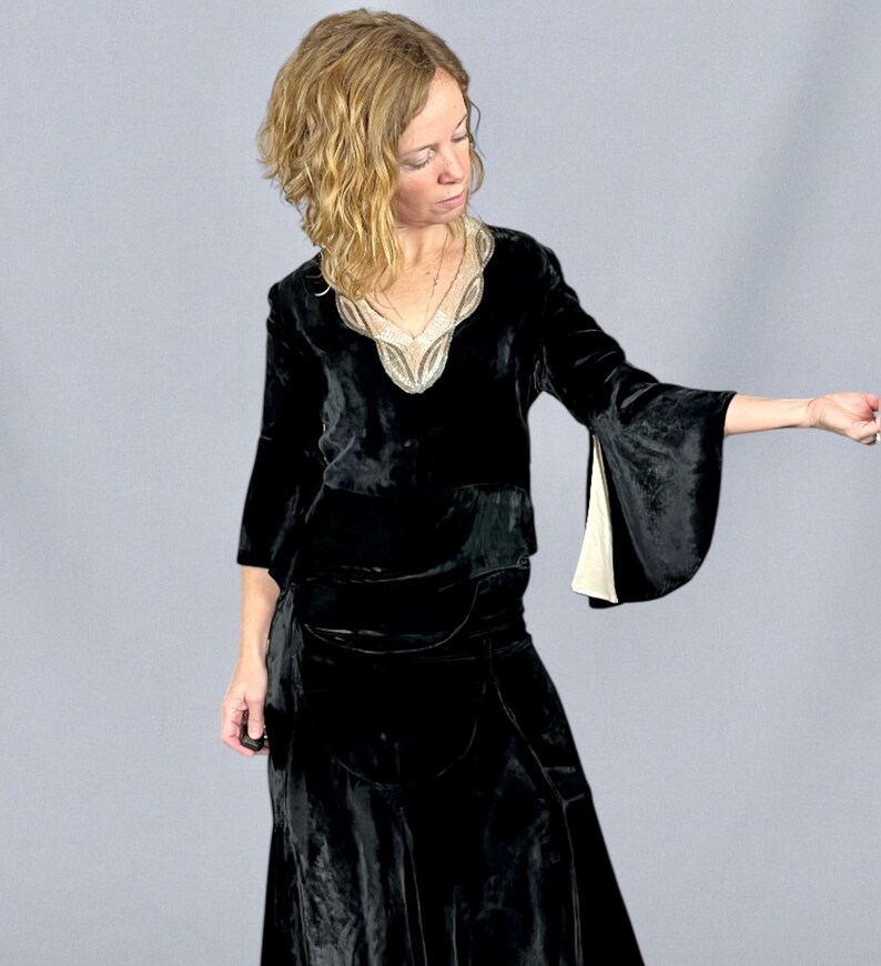 Puede incluir: Un vestido de terciopelo negro con escote en V y mangas acampanadas. El escote est&aacute; adornado con un ribete decorativo de color claro. El vestido parece ser de estilo vintage o retro, con una falda larga y un corpi&ntilde;o ajustado. Las mangas tienen un forro crema en contraste.