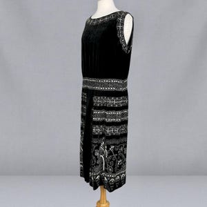 May include: A sleeveless black velvet dress with intricate silver-colored embroidery. The dress features detailed patterns around the neckline, waist, and hem. The dress is displayed on a wooden mannequin, showcasing its vintage design.