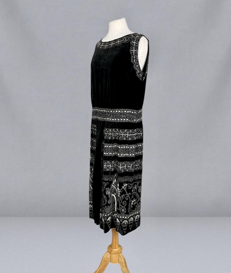 May include: A sleeveless black velvet dress with intricate silver-colored embroidery. The dress features detailed patterns around the neckline, waist, and hem. The dress is displayed on a wooden mannequin, showcasing its vintage design.