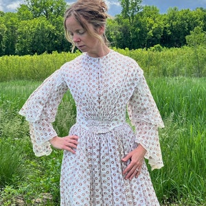 May include: A white dress with a brown floral pattern and a gathered waist. The dress has long sleeves with ruffles at the cuffs. The model is standing in a field of green grass.