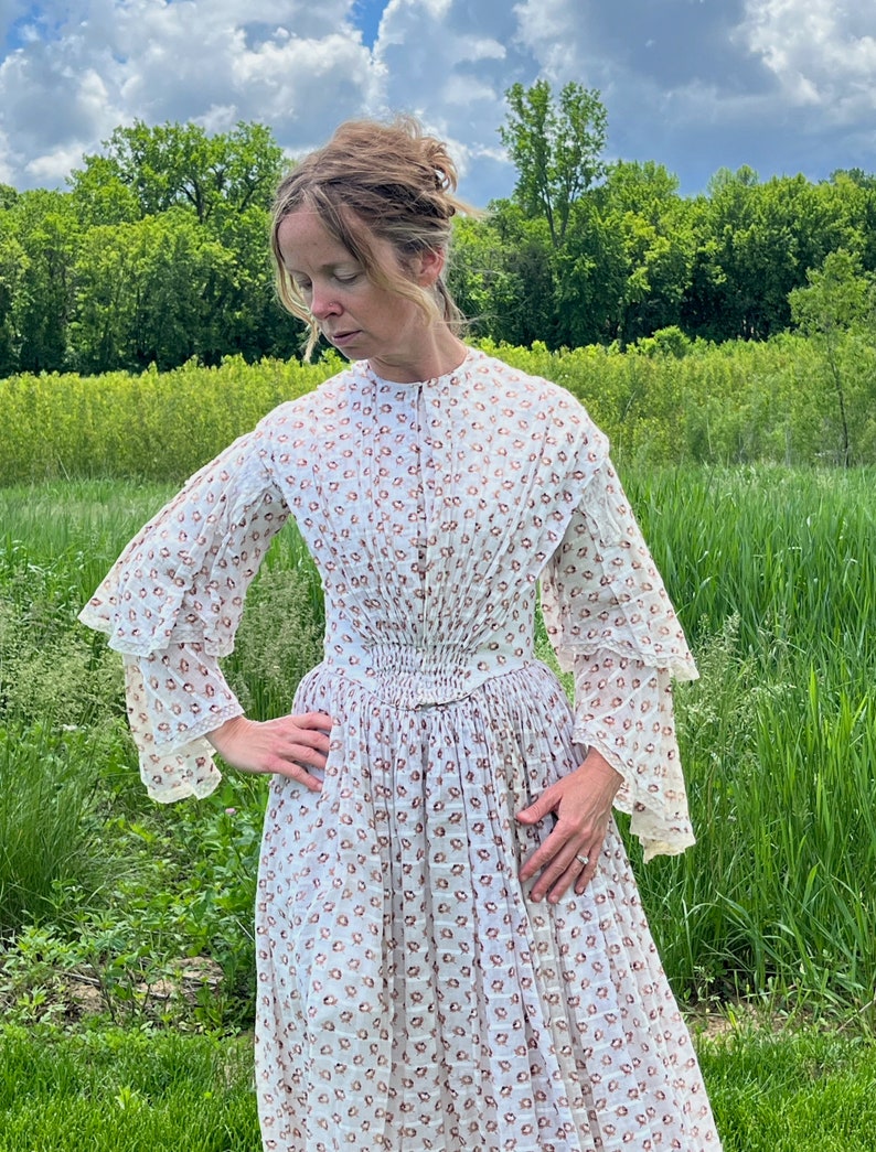 May include: A white dress with a brown floral pattern and a gathered waist. The dress has long sleeves with ruffles at the cuffs. The model is standing in a field of green grass.