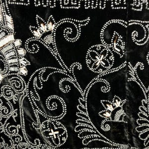 May include: Close-up of a black velvet textile with intricate silver beadwork. The design features floral and geometric patterns, with beaded borders and swirling vines. The beads are small and create a shimmering effect.