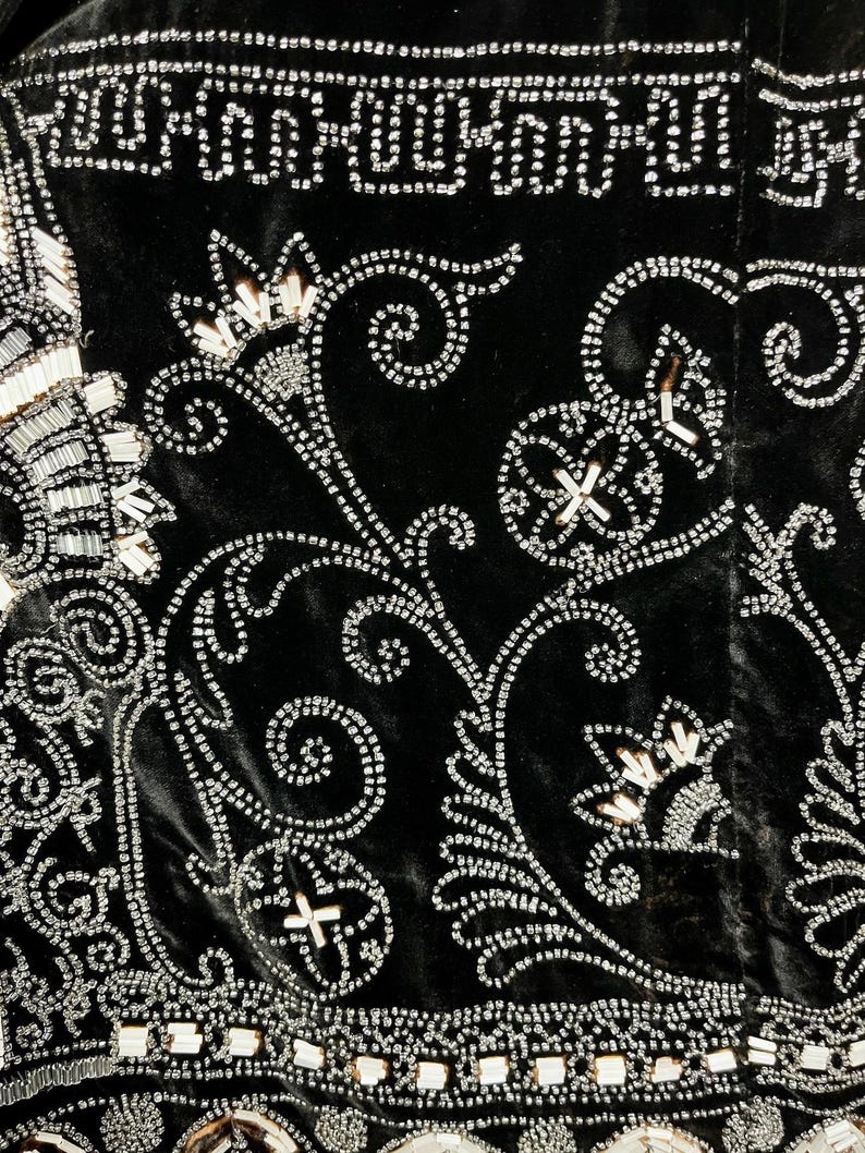 May include: Close-up of a black velvet textile with intricate silver beadwork. The design features floral and geometric patterns, with beaded borders and swirling vines. The beads are small and create a shimmering effect.