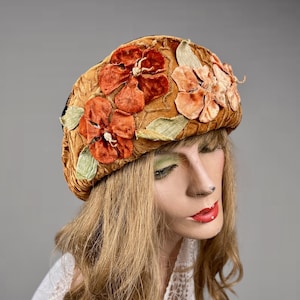 May include: A vintage-style hat with a velvet texture, adorned with orange and peach-colored fabric flowers and green leaves. The hat has a rounded shape and is displayed on a mannequin head. The hat's color is a warm, golden brown.