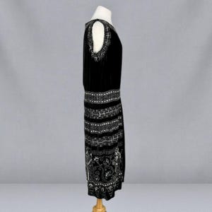 May include: A black velvet dress with white beaded embellishments. The dress features a drop waist and a knee-length hemline. The shoulders have cut-out details with decorative beading. The dress is displayed on a mannequin.