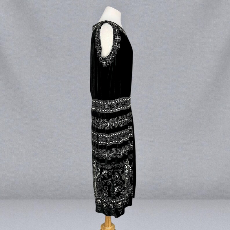 May include: A black velvet dress with white beaded embellishments. The dress features a drop waist and a knee-length hemline. The shoulders have cut-out details with decorative beading. The dress is displayed on a mannequin.